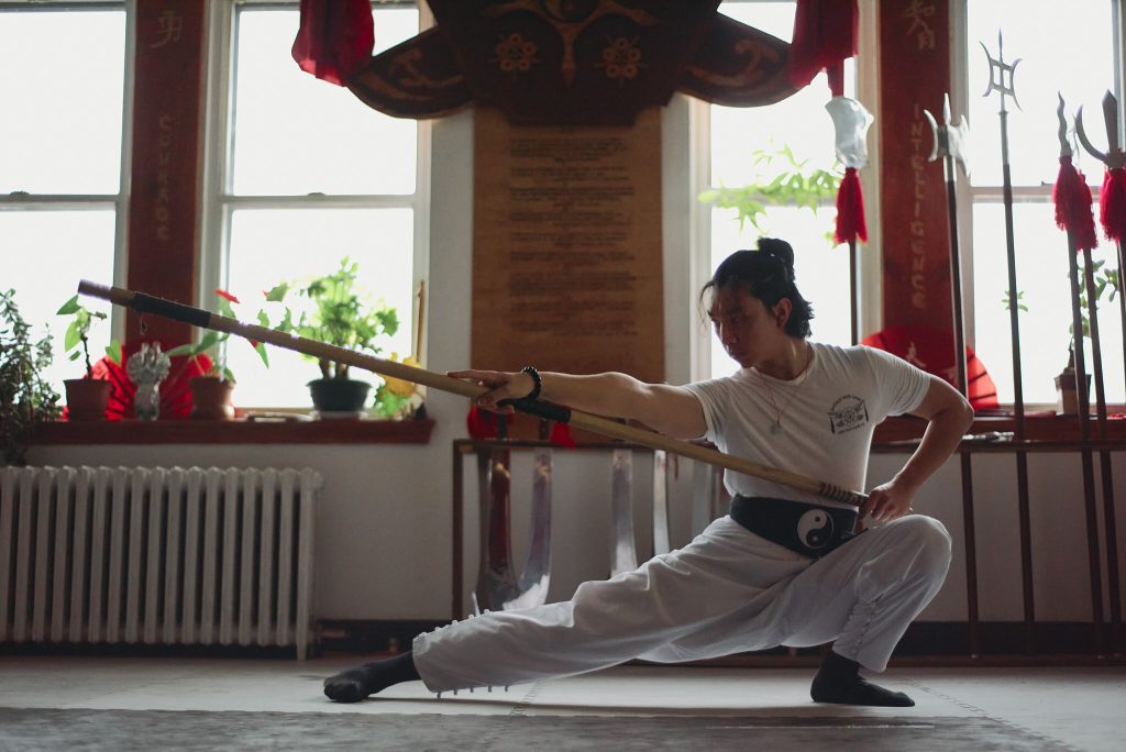 Kung Fu practitioner in a low stance holding a long staff, focusing intently in a traditional martial arts studio with plants and weapons displayed in the background.