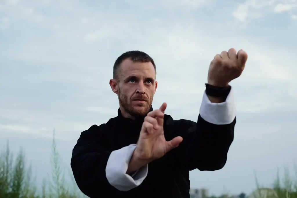 A martial artist in traditional black and white Kung Fu attire practicing focused hand techniques outdoors under a cloudy sky.