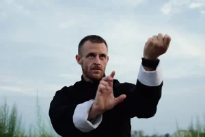 A martial artist in traditional black and white Kung Fu attire practicing focused hand techniques outdoors under a cloudy sky.